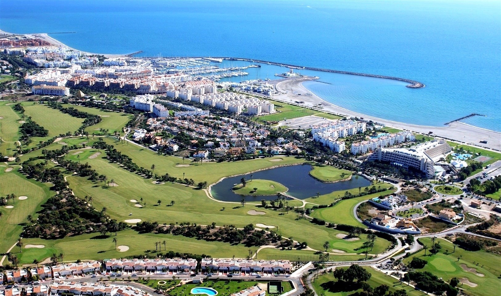 Vista aérea de una ciudad costera con un campo de golf, un puerto deportivo y playas a orillas del mar azul.