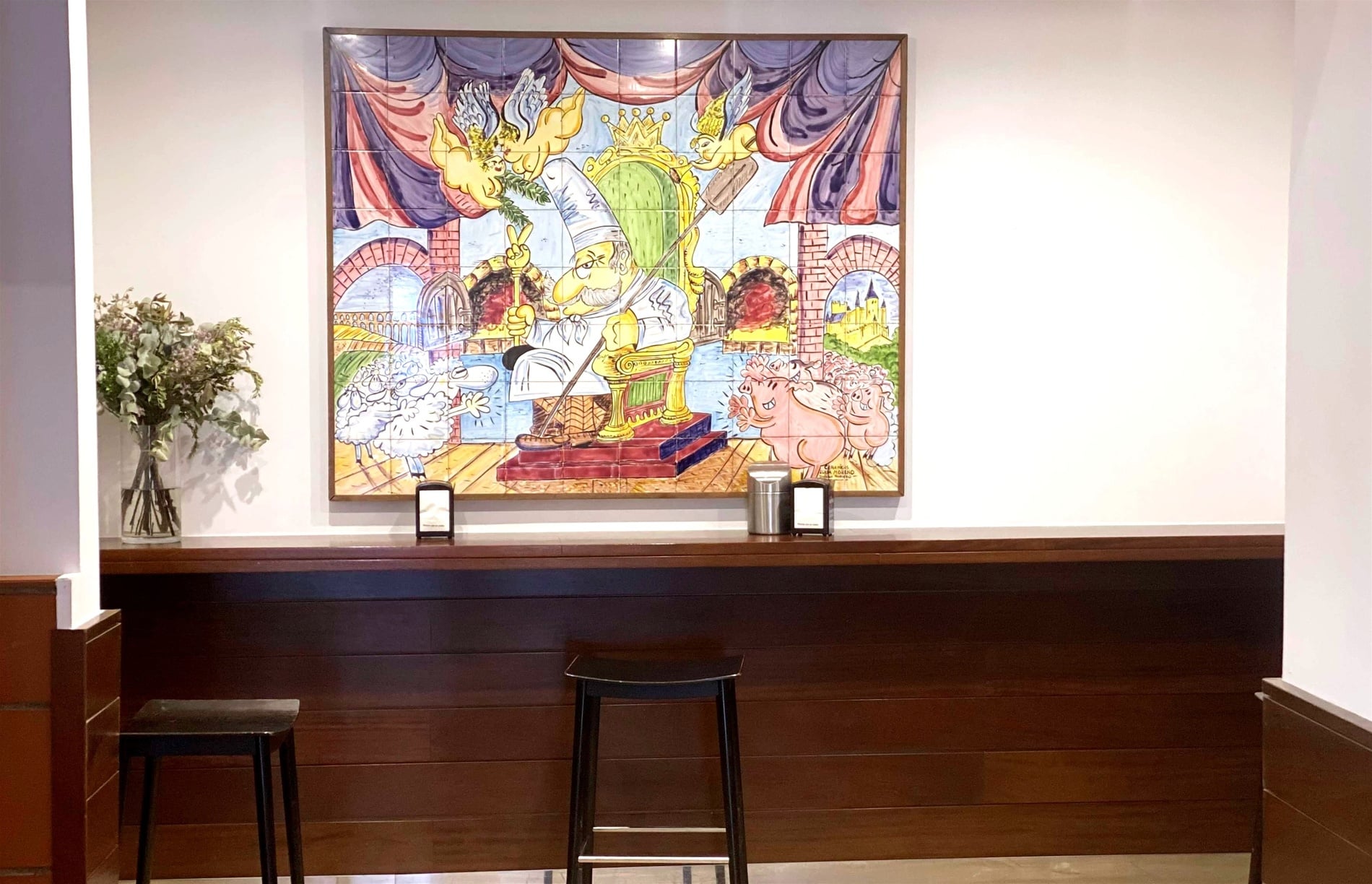 A large, colorful tile mural depicting a chef on a golden throne surrounded by cherubs, sheep, and pigs is displayed above a wooden counter with two bar stools.