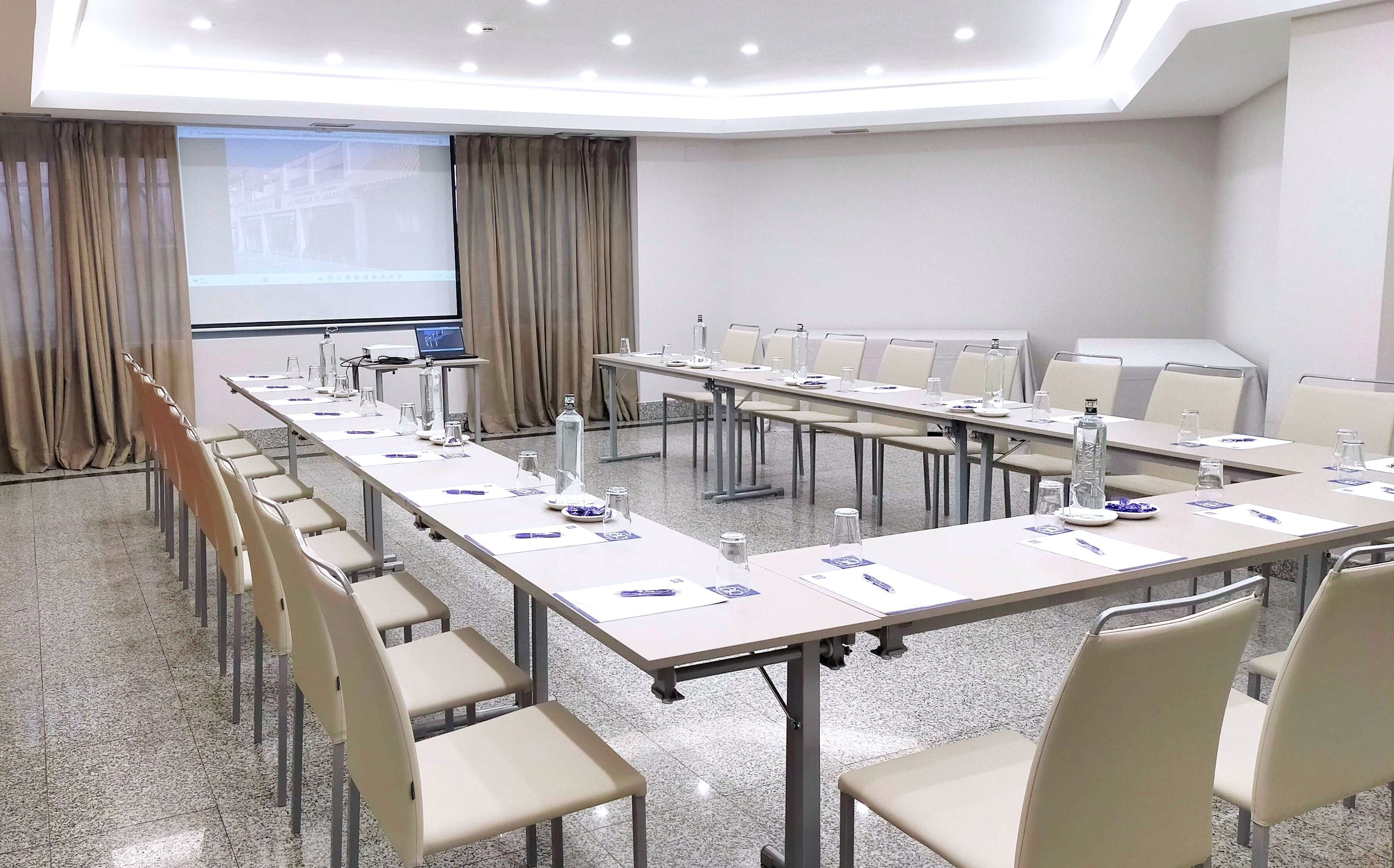 A brightly lit conference room is arranged with tables in a U-shape, set with water bottles, glasses, and notepads, and features a projector screen displaying an image on the left wall.