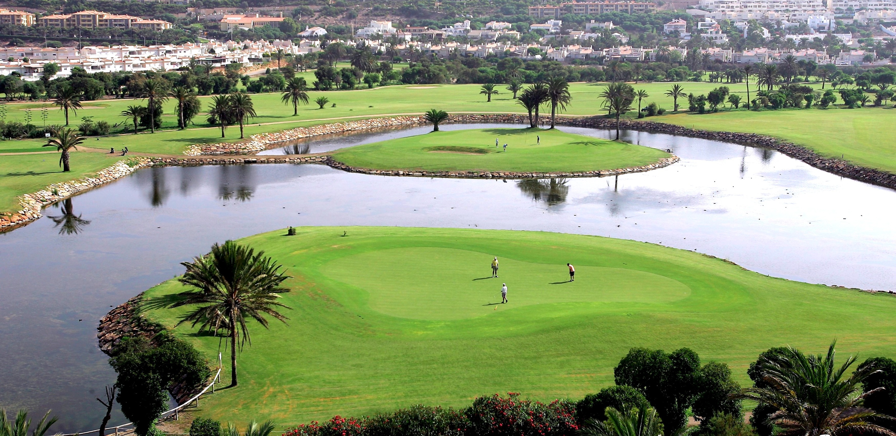 An elevated view shows a lush green golf course with water features, island greens, and palm trees, overlooked by residential buildings on a distant hillside.