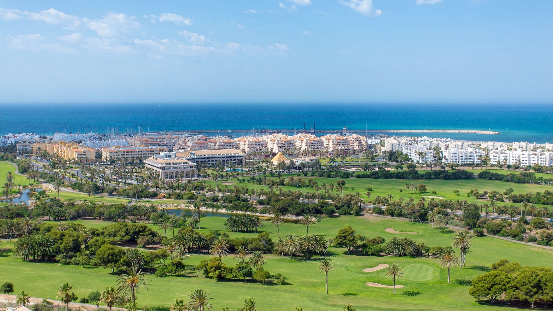 Vista panorámica de un complejo turístico costero que incluye un campo de golf, edificios, un puerto deportivo con barcos y el mar azul bajo un cielo despejado.