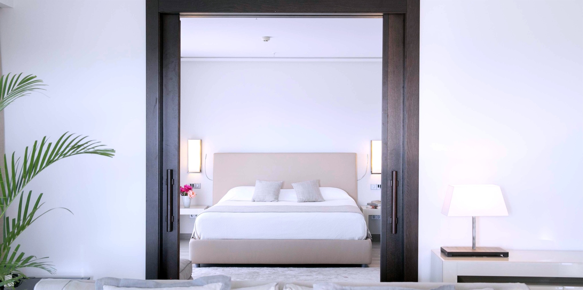 La imagen muestra un dormitorio moderno con una cama grande y mesitas de noche, visto a través de una puerta corrediza de madera oscura desde una sala de estar con un sofá.