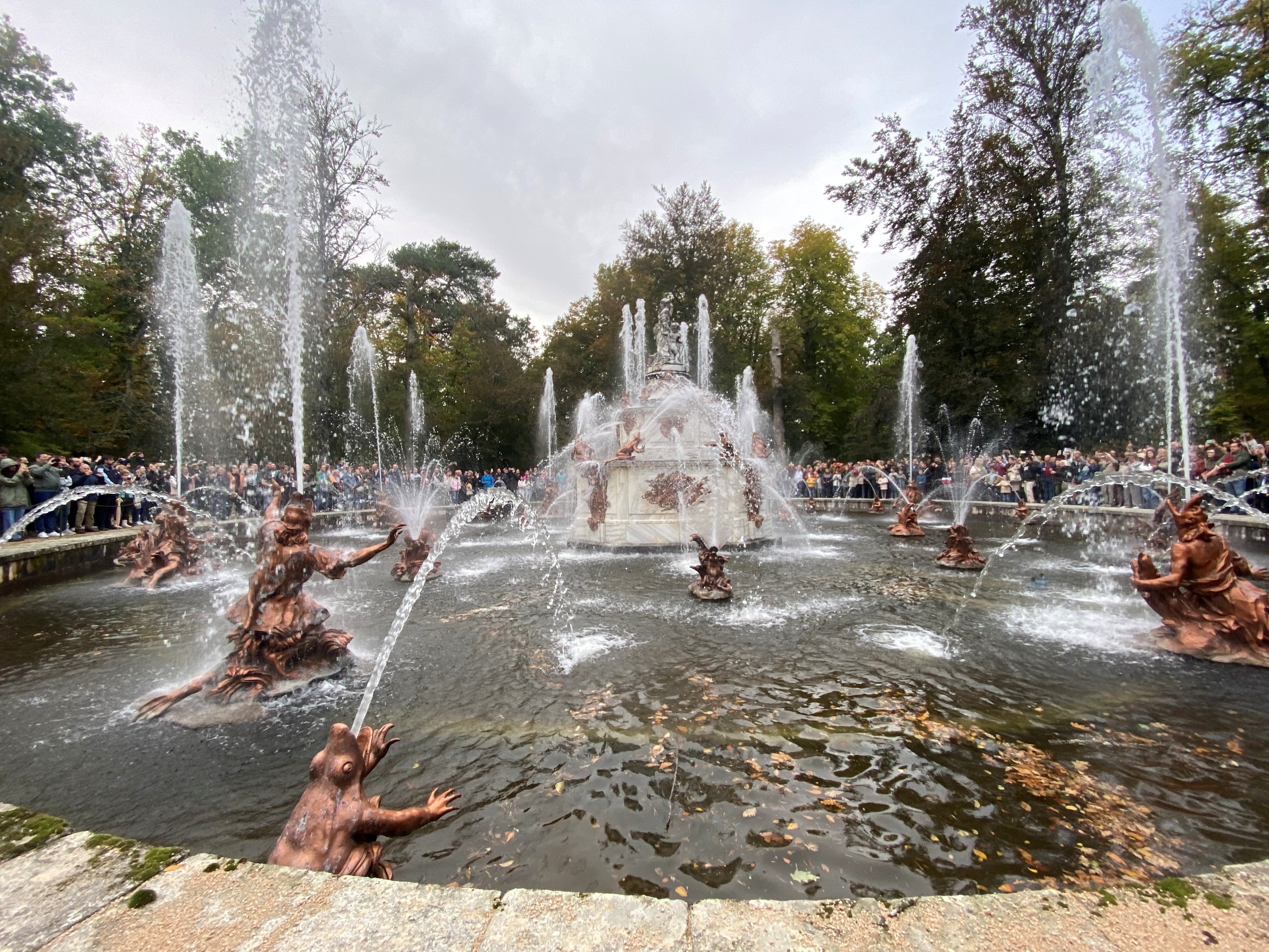 A grand, ornate fountain featuring multiple water jets and bronze statues of mythological figures, surrounded by a large crowd of onlookers and lush trees under an overcast sky.