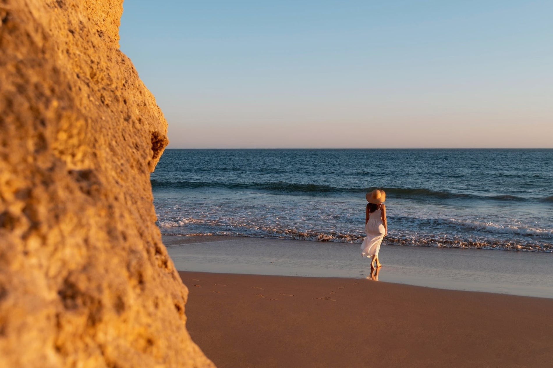 Una mujer con un vestido blanco y sombrero camina por una playa arenosa junto a un acantilado rocoso, con el océano de fondo al atardecer.