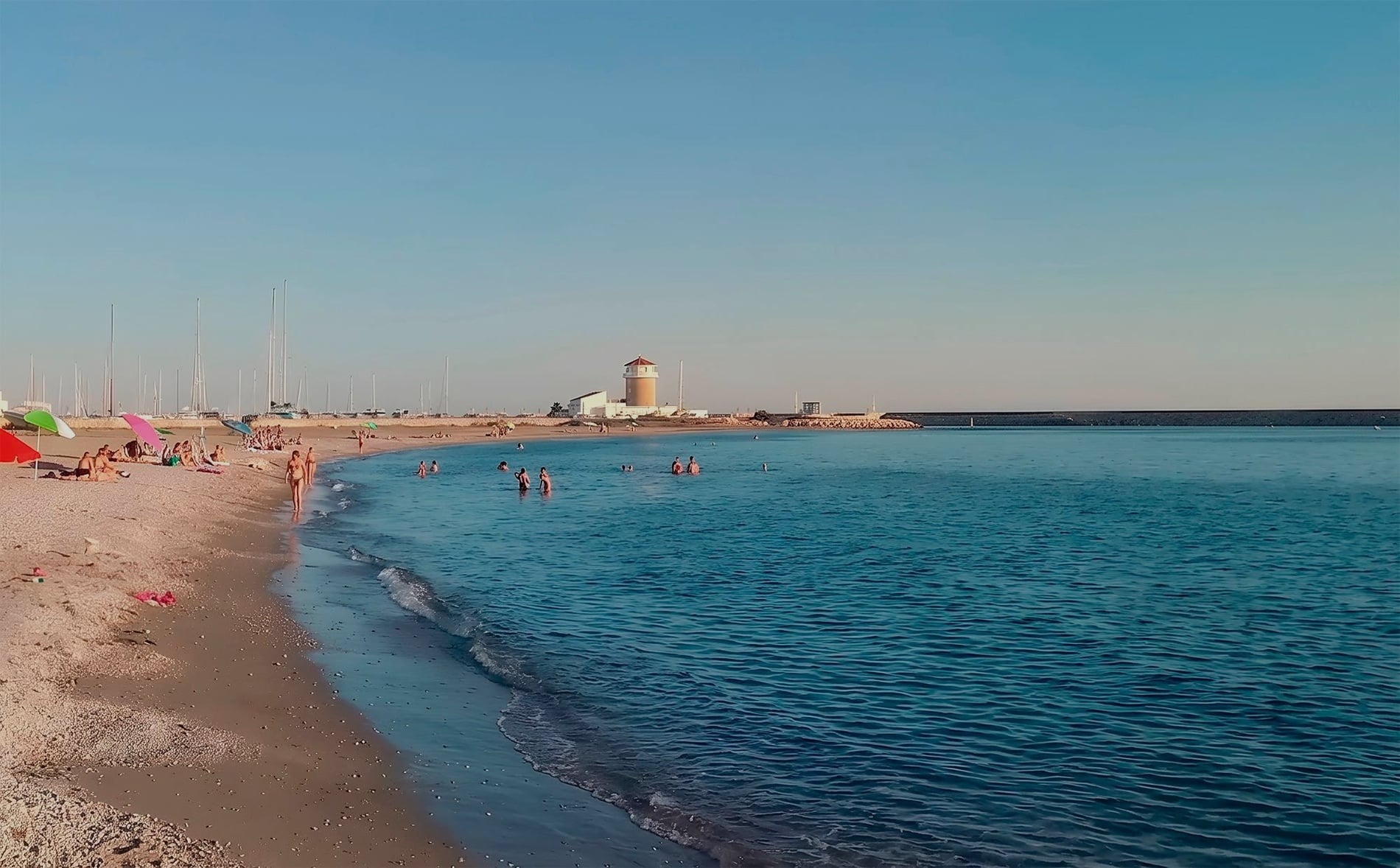 Playa en AR: Golf Almerimar, gente nadando y veleros, con un faro al fondo.