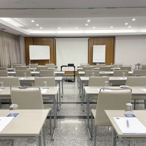 Una sala de conferencias o formación vacía y preparada, con filas de mesas y sillas individuales, una pantalla de proyector y pizarras blancas.
