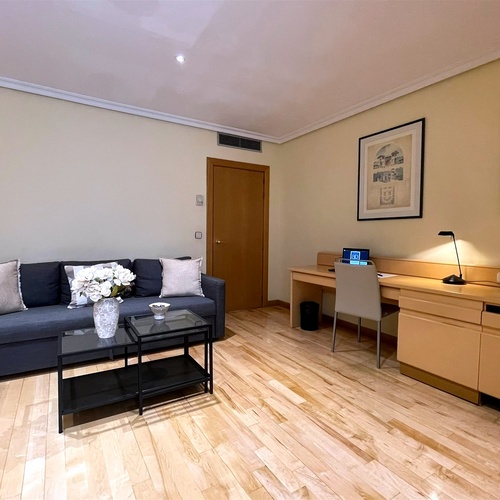 Una habitación amueblada con un sofá gris, un escritorio de madera, mesas auxiliares y cuadros decorativos, todo sobre un suelo de madera clara.