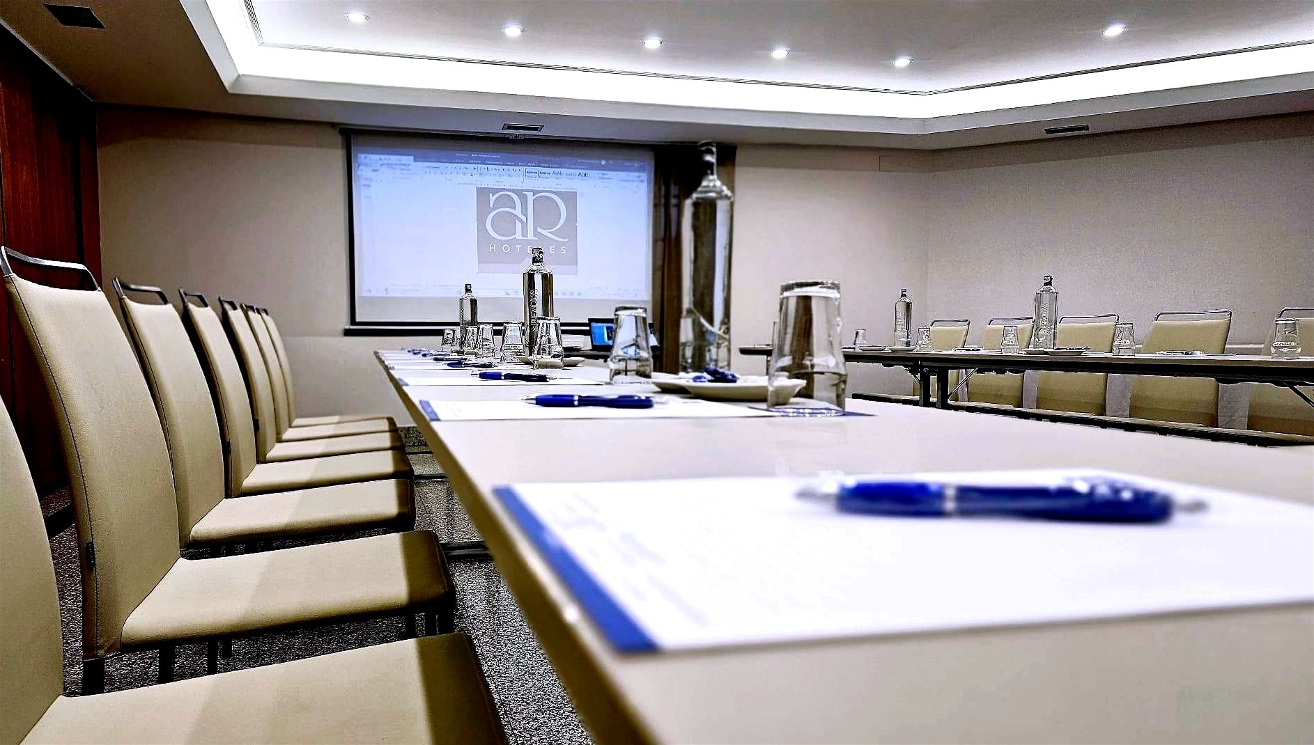 Una sala de reuniones moderna y bien equipada, con mesas dispuestas en forma de "U" o rectángulo, sillas, botellas de agua, vasos y material de escritura, y una pantalla de proyección al fondo mostrando el logo "AR HOTELS".
