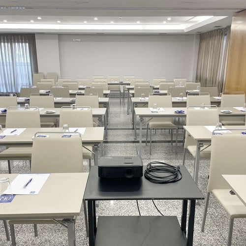 Una sala de conferencias vacía con múltiples filas de mesas y sillas preparadas para un evento, y un proyector en primer plano.