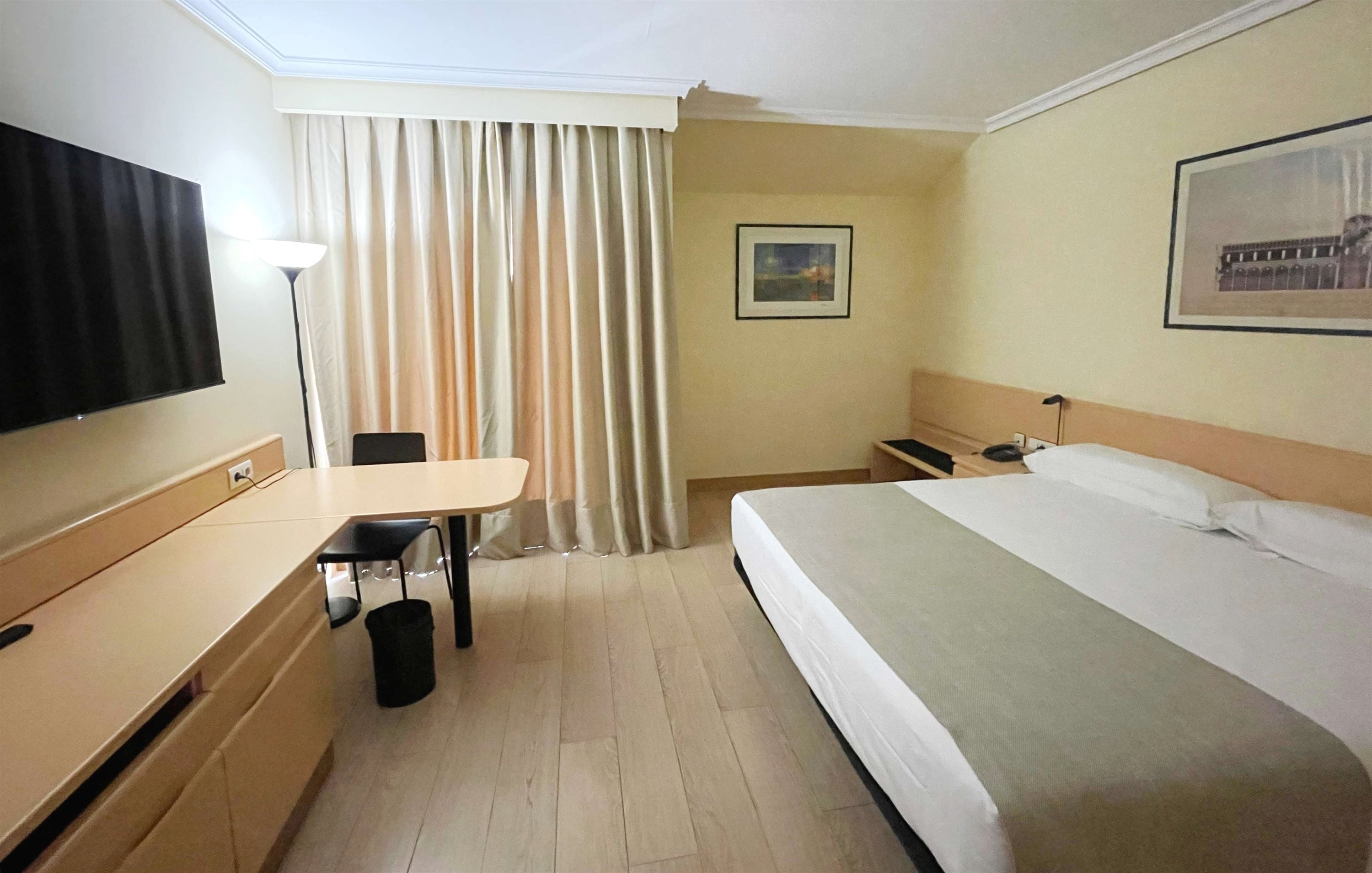 La imagen muestra una habitación de hotel sencilla y luminosa con una cama hecha, dos cuadros en la pared y cortinas de color beige.
