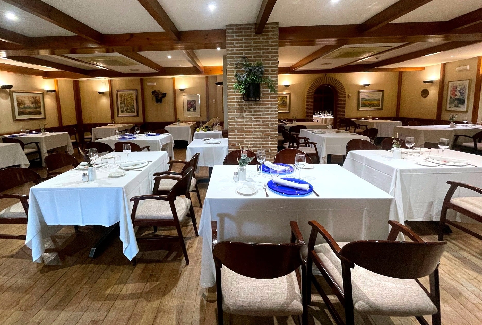 This image captures a well-lit, empty restaurant dining room featuring numerous tables dressed with white tablecloths and place settings, dark wooden chairs, a wood-beamed ceiling, and artwork on the walls.