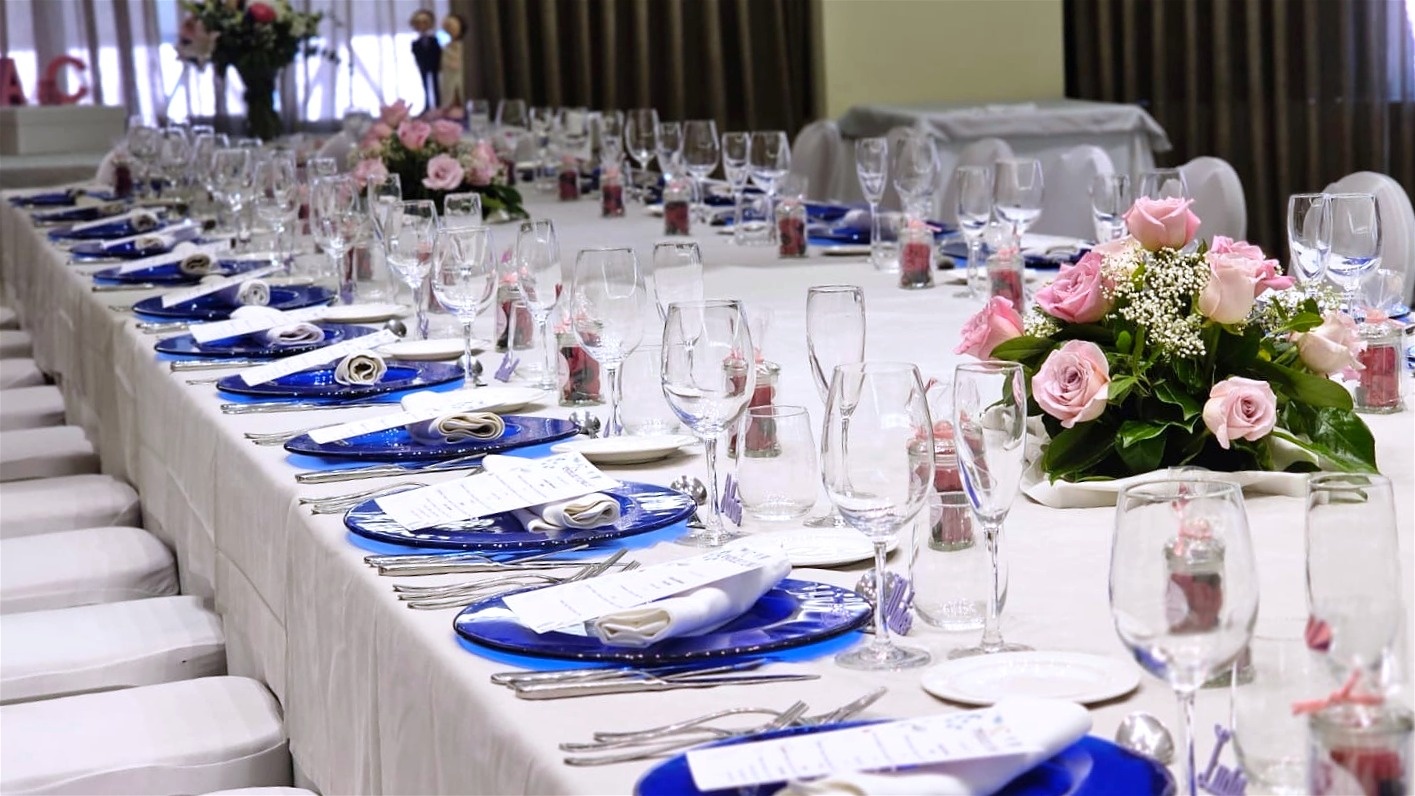 Una larga mesa elegantemente dispuesta para un evento presenta manteles blancos, platos azules vibrantes, copas variadas y delicados centros de mesa de rosas rosadas.