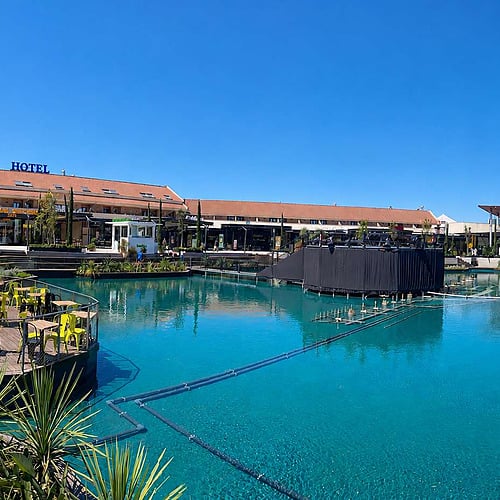 Outdoor shopping center with a turquoise water feature, walkways, and buildings. Explore AR: Parquesur shopping mall.