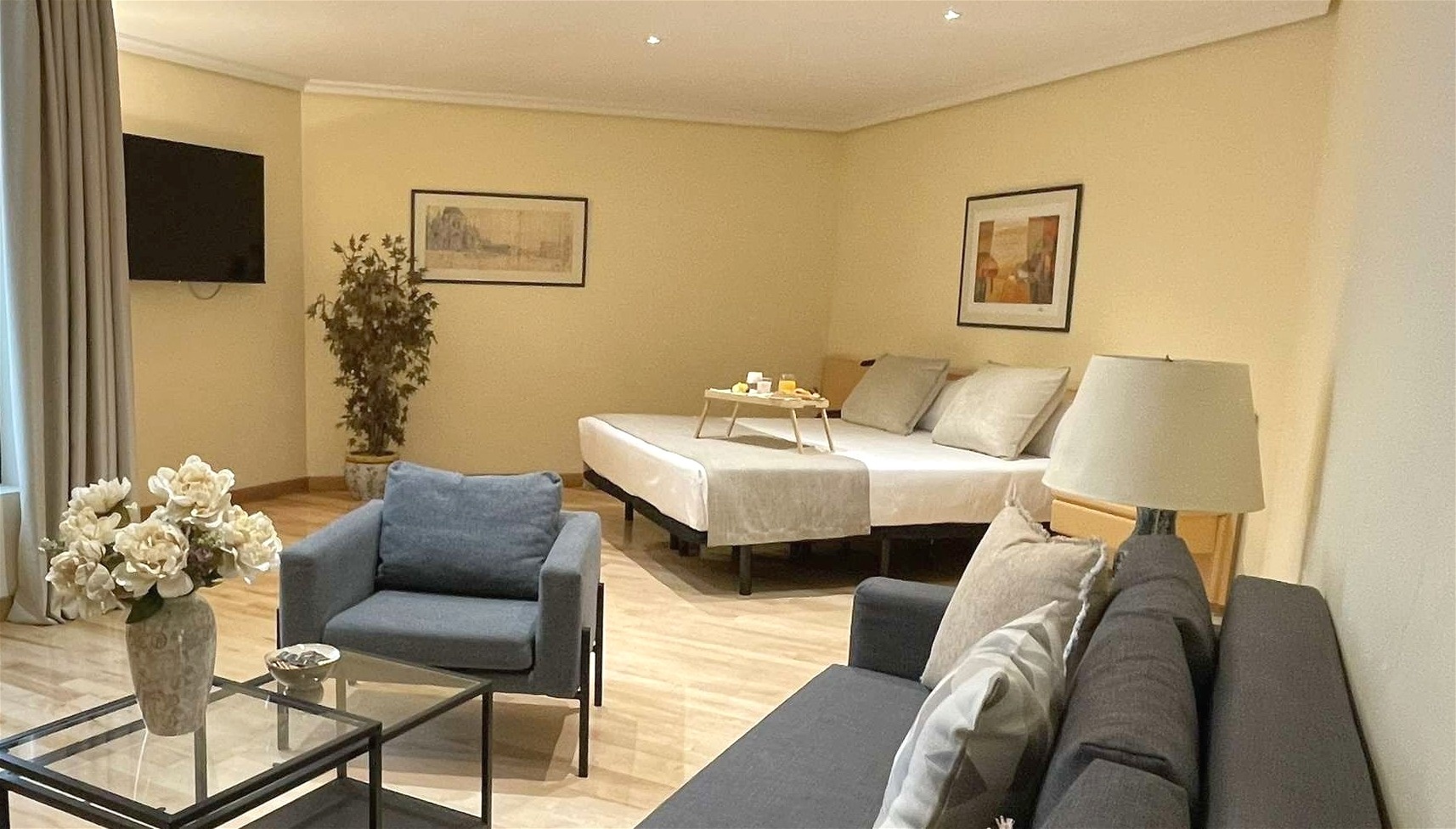 Un acogedor apartamento tipo estudio presenta paredes color crema, suelo de madera, un sofá y un sillón grises, una cama con una bandeja de desayuno, una televisión montada en la pared y una ventana grande.