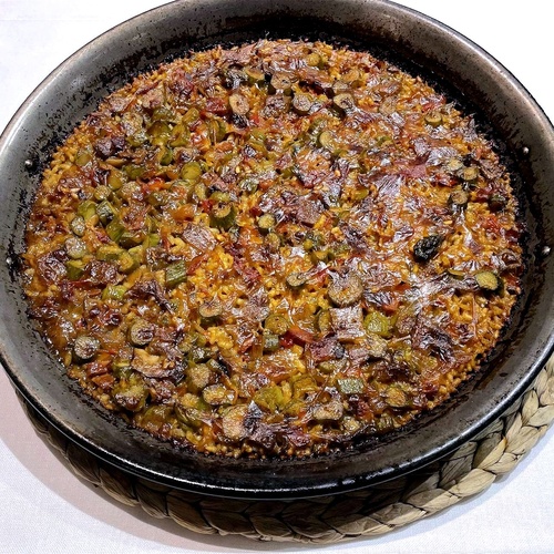 Una paellera grande llena de arroz con verduras verdes y trozos oscuros, apoyada sobre un salvamanteles de mimbre en un mantel blanco.