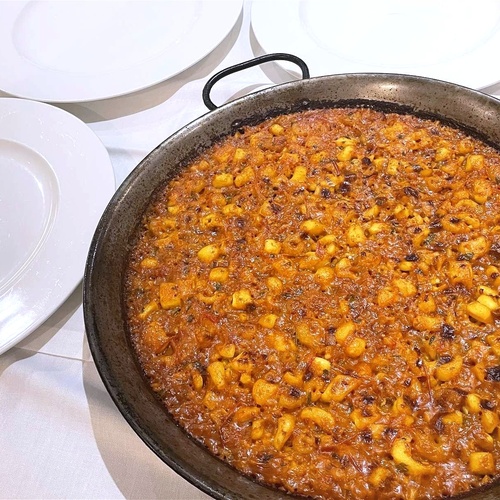 En la imagen se ve una paellera grande llena de un plato de fideuá o similar, con trozos de pasta y un sofrito rojizo, sobre un mantel blanco junto a varios platos vacíos.