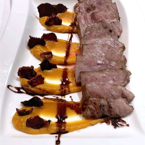 En un plato blanco se presentan lonchas de carne con sal gruesa junto a porciones de puré anaranjado, decoradas con trozos oscuros crujientes y un glaseado.