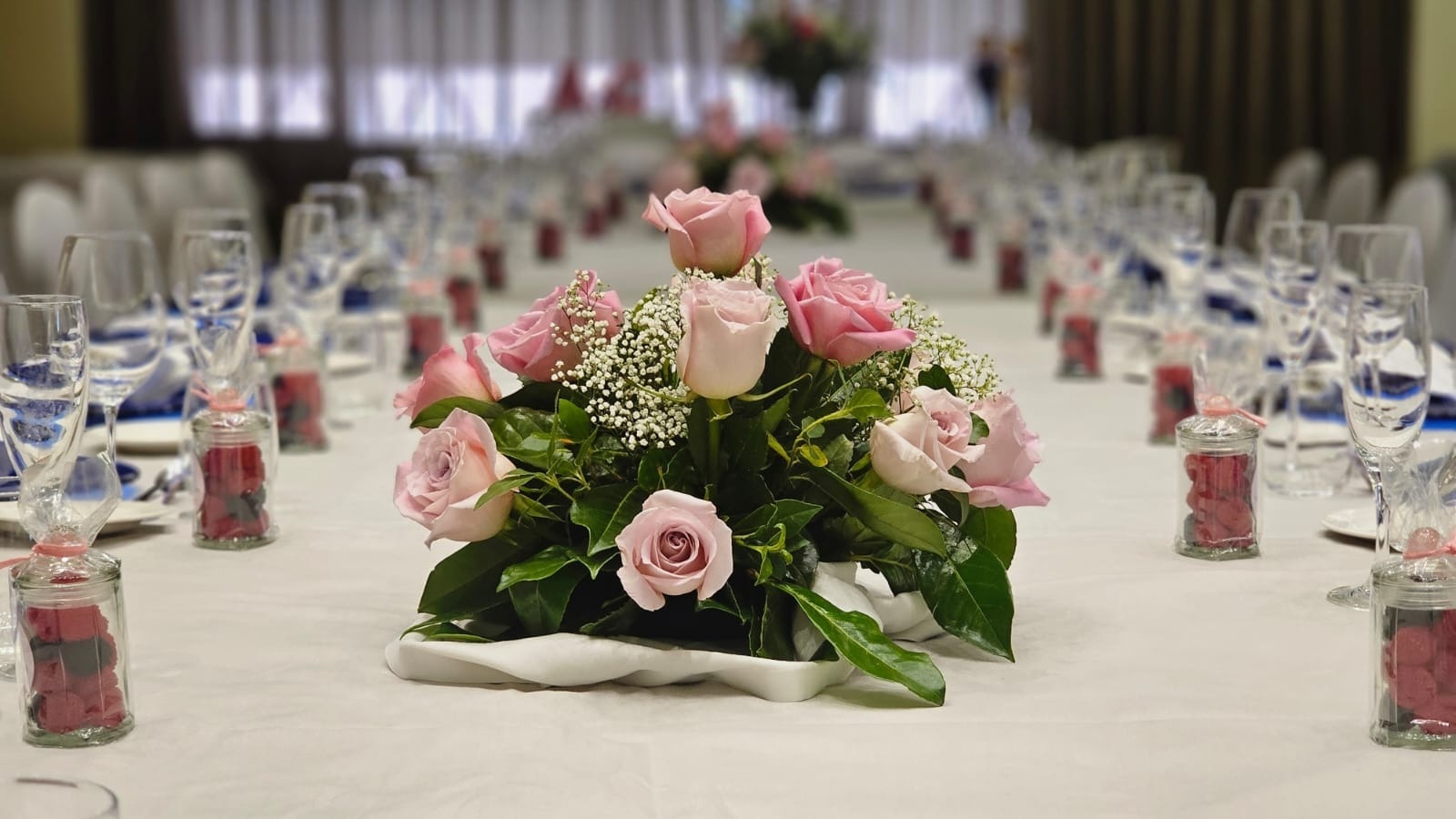 Una mesa larga, cubierta con un mantel blanco, está adornada con un centro de rosas rosadas, copas y pequeños frascos con golosinas rojas, dispuesta para un evento.