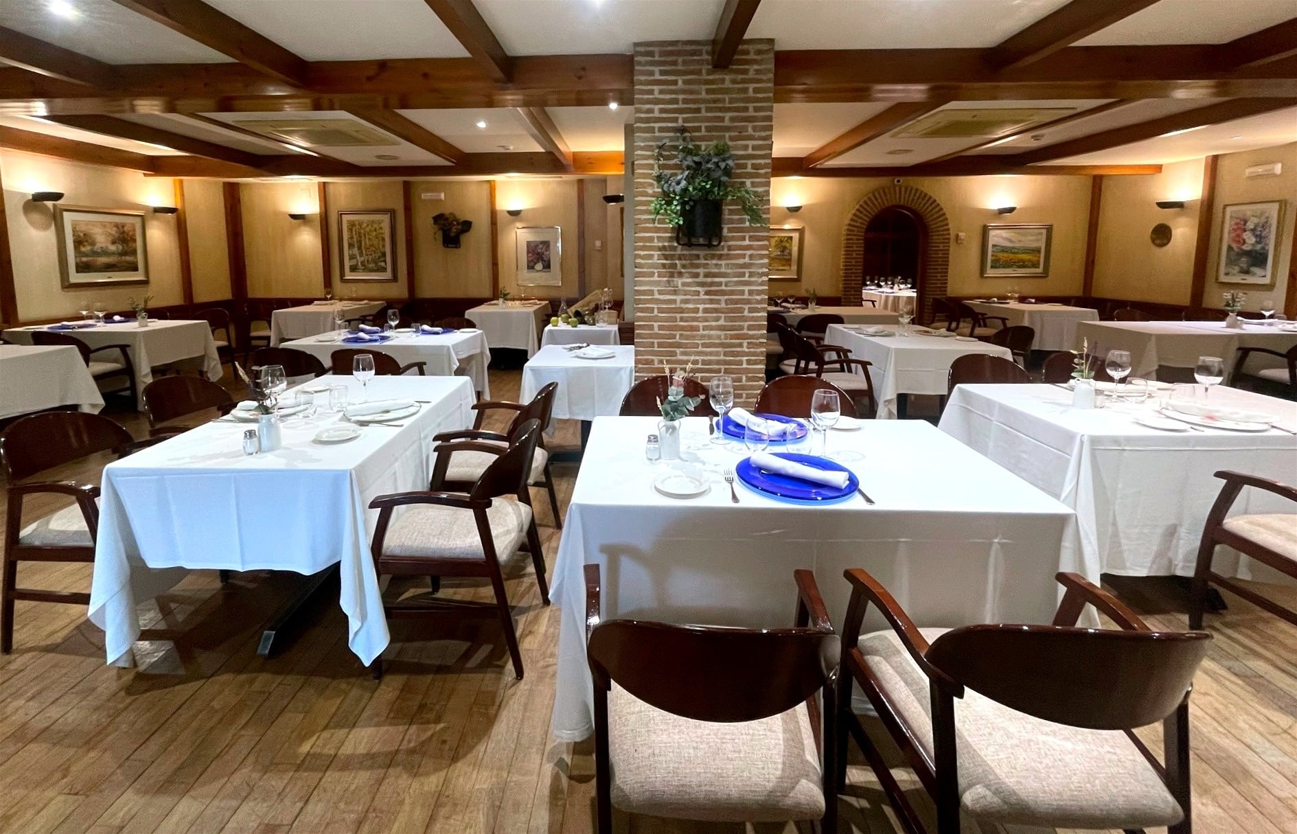 La imagen muestra el interior de un restaurante elegante con mesas vestidas con manteles blancos, sillas de madera, un suelo de parqué, vigas de madera en el techo y decoraciones en las paredes.