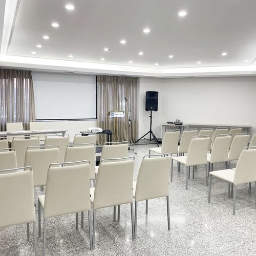 Una sala de conferencias se muestra preparada para un evento, equipada con filas de sillas, un podio, proyector con pantalla y un altavoz, bajo un techo falso con iluminación.