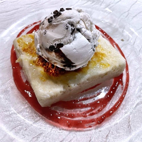 Un postre con una porción rectangular de tarta o bizcocho, helado de vainilla con trocitos de chocolate, y salsa roja en un plato de cristal texturizado.