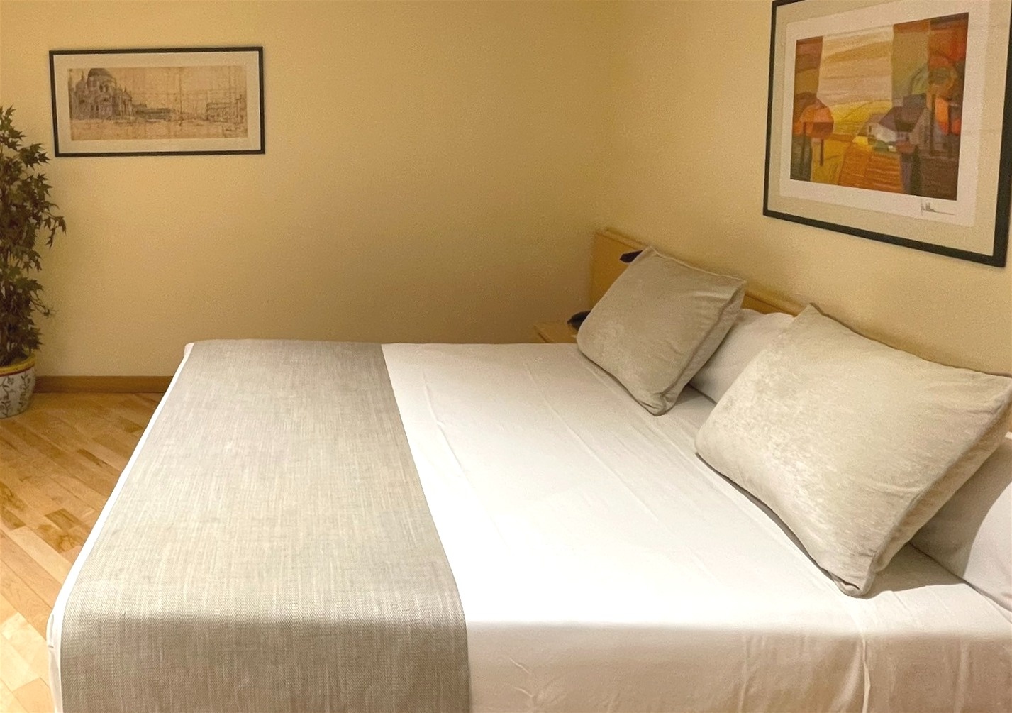 This image captures a bright and spacious studio apartment, featuring a bed, a sofa with throw pillows, armchairs, a coffee table with a laptop, and a flat-screen TV against warm yellow walls and light wooden flooring.