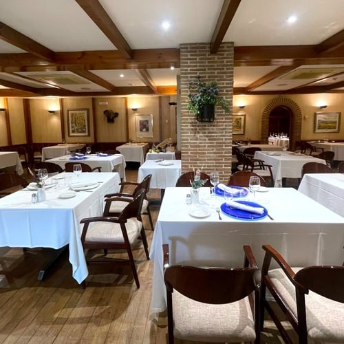 This image captures the inviting interior of a restaurant featuring multiple dining tables set with white tablecloths, dark wooden chairs, and place settings, all under a ceiling with prominent wooden beams and soft lighting.