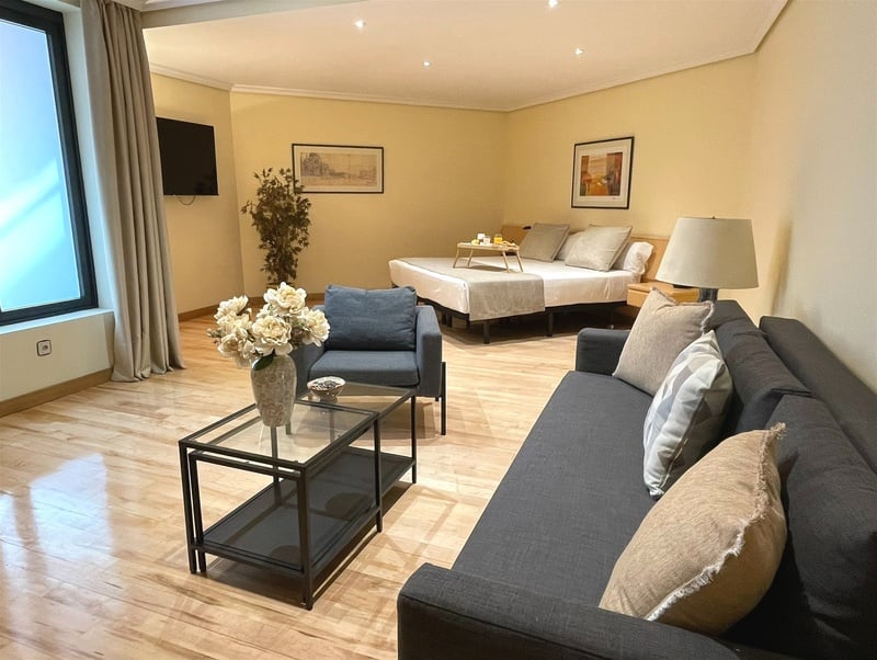 A bright, open-plan studio room features a dark grey sofa and armchair, a glass coffee table, and a bed with a breakfast tray, all illuminated by a large window and recessed lighting against light yellow walls and wooden floors.