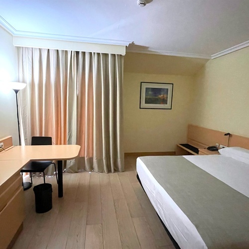 La imagen muestra una habitación de hotel con una cama, un escritorio con televisión, cortinas beige y un suelo de madera clara.