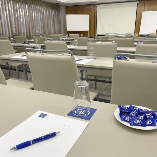 Una sala de conferencias está preparada con mesas y sillas dispuestas en filas, cada puesto con material de escritura, un vaso y dulces, y con una pantalla de proyección y una pizarra al frente.
