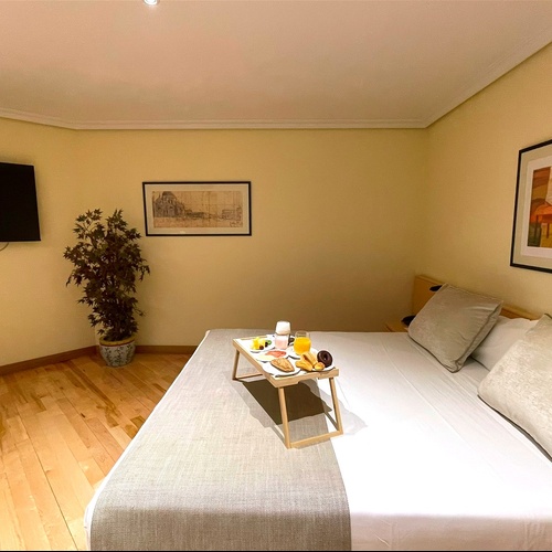 Una acogedora habitación de hotel se muestra con una cama doble, un televisor en la pared, un desayuno en bandeja y decoraciones.