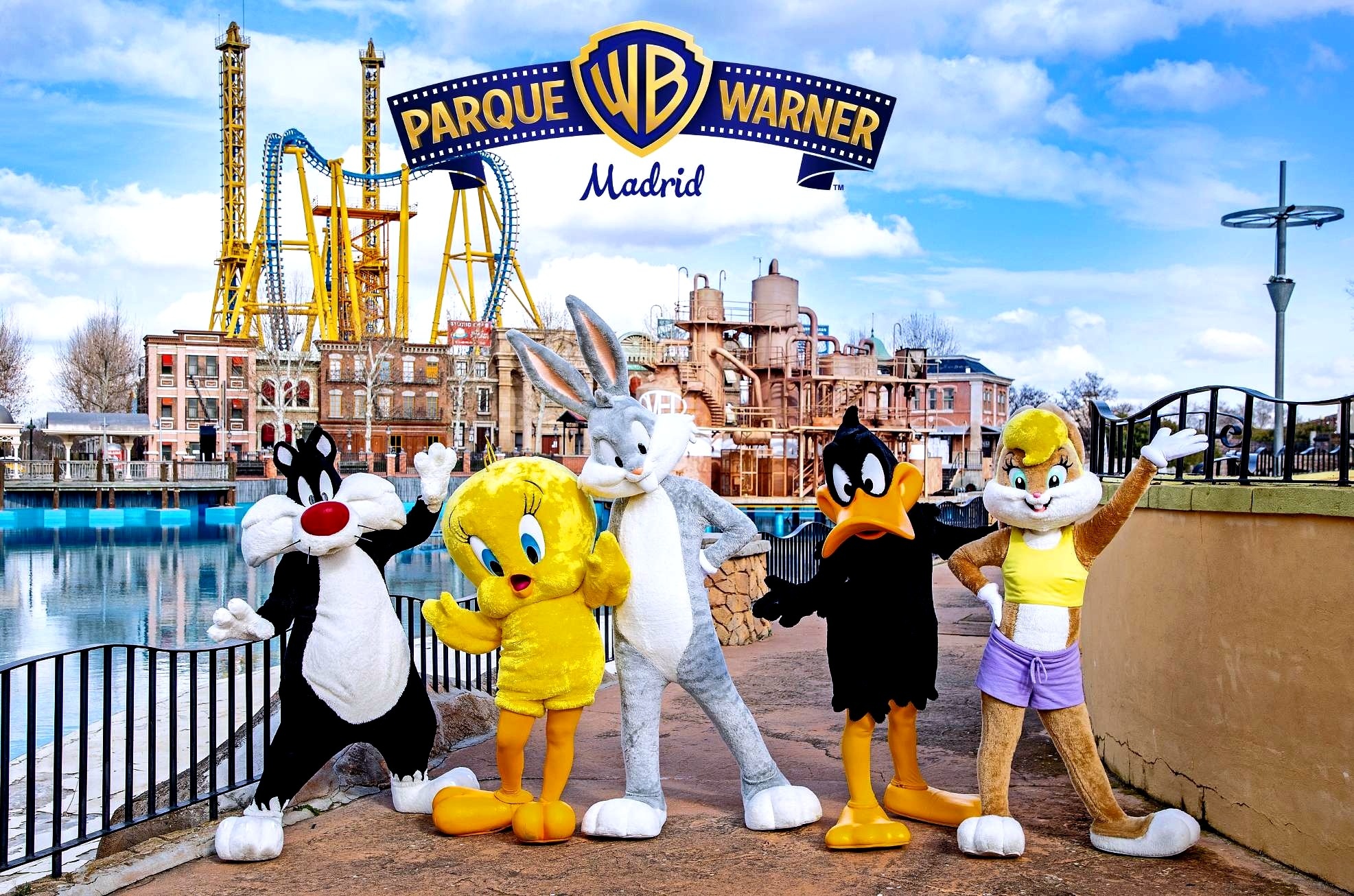 Five Looney Tunes characters in mascot costumes (Sylvester, Tweety, Bugs Bunny, Daffy Duck, and Lola Bunny) pose in front of the Parque Warner Madrid theme park, featuring a roller coaster and buildings, under a cloudy sky.