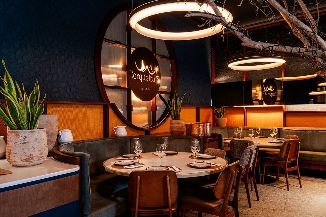 A imagem retrata um restaurante moderno e acolhedor, com mesas postas, plantas e iluminação circular, destacando o logótipo 'Cerqueira's' e um interior com paredes texturizadas e detalhes em madeira e tons alaranjados.