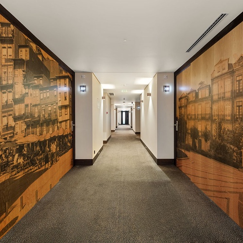 Um corredor moderno de hotel apresenta paredes decoradas com grandes murais em tom sépia de cenas urbanas antigas, com números de quarto iluminados visíveis.