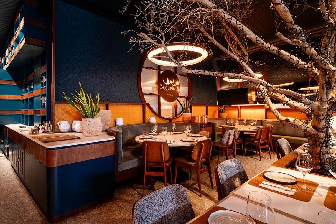 O interior de um restaurante moderno e elegante, com predominância de azul escuro e detalhes em laranja e madeira, é realçado por uma grande árvore seca decorada com luzes e mesas postas.