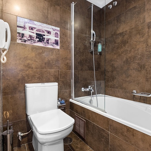 Um banheiro compacto com paredes e chão de azulejos marrons, equipado com um vaso sanitário branco, uma banheira com chuveiro e divisória de vidro, um telefone fixo na parede e um quadro decorativo.