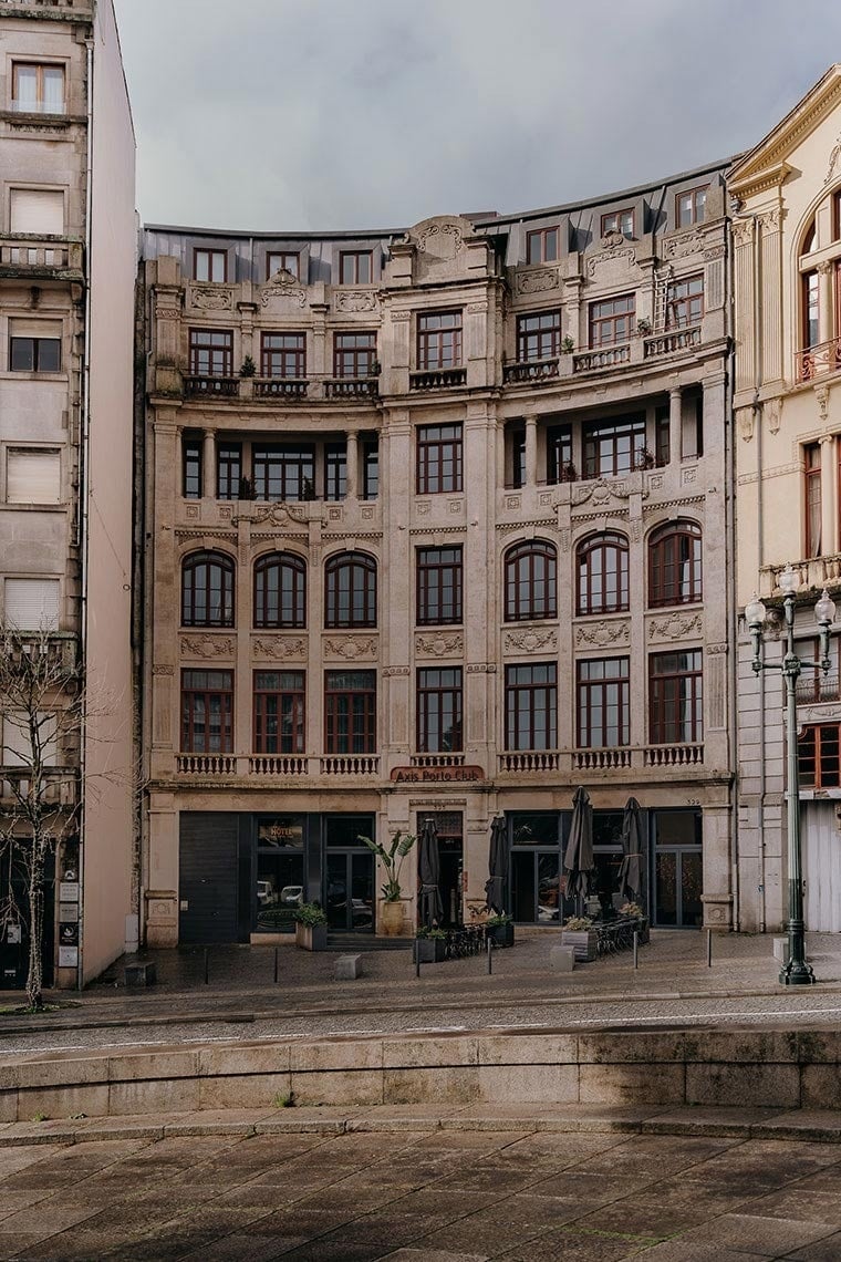 Um grande edifício de pedra com uma fachada curva ornamentada e janelas de madeira escura, apresentando lojas no piso térreo e a inscrição 'Axis Porto Club', sob um céu nublado.