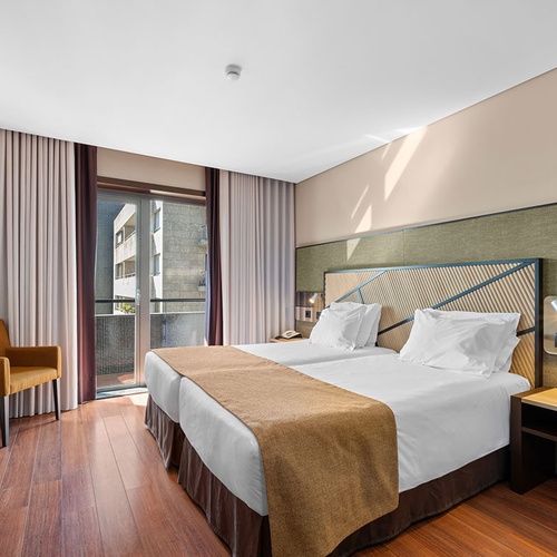 Um quarto de hotel moderno com duas camas individuais, chão de madeira, uma secretária e acesso a uma varanda, predominantemente em tons neutros.