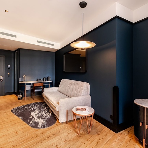 A stylish room features dark blue walls, light hardwood flooring, a grey sofa, a compact workspace, and a sleek pendant light.
