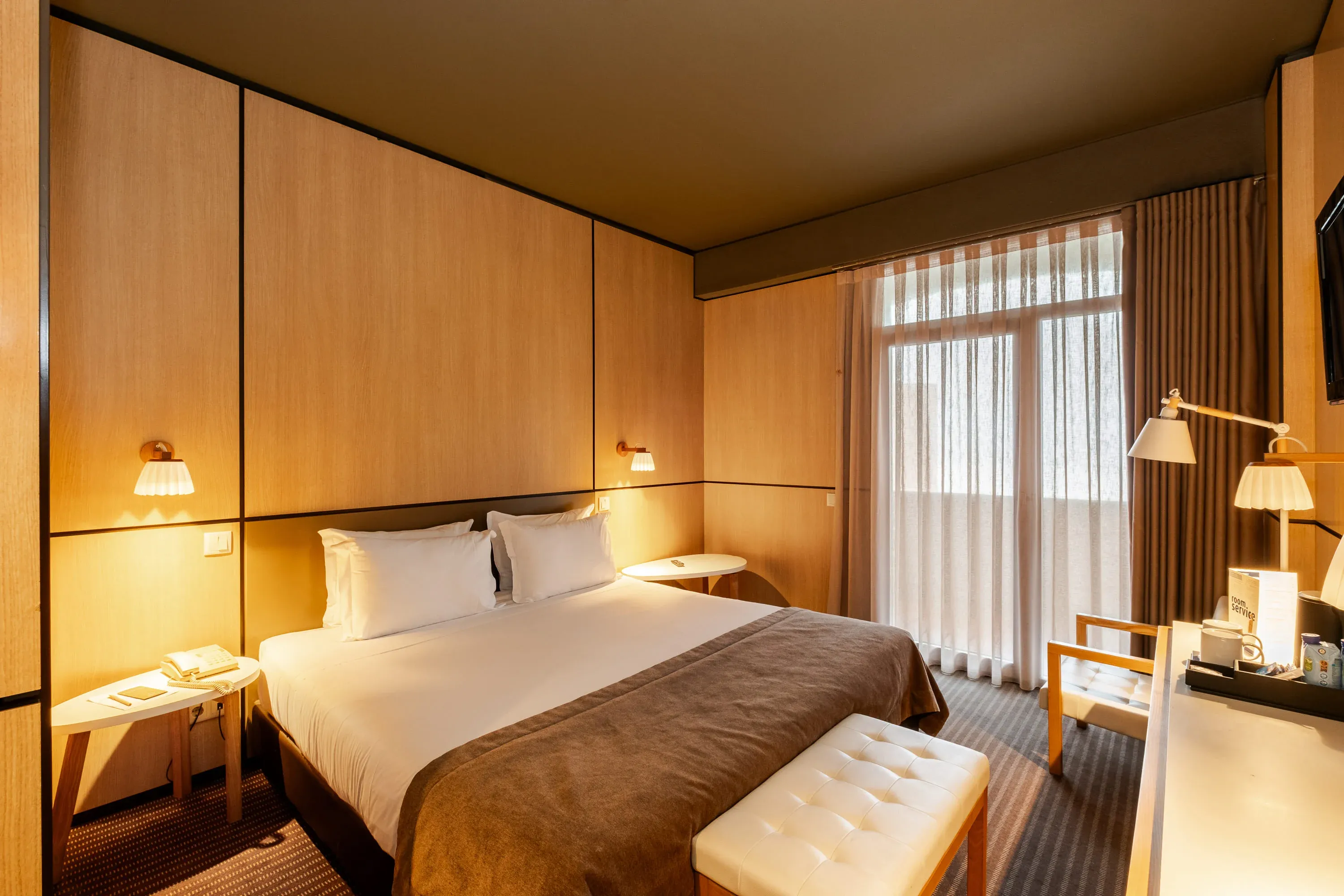 Quarto de hotel moderno com cama confortável, mesa de cabeceira e janela com cortinas leves. Vermar | Axis Hotéis & Golfe | Website Oficial