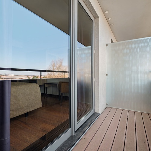 Uma varanda com piso de deck de madeira e uma porta de correr de vidro que reflete o interior, ladeada por uma divisória de vidro fosco e com vista para o exterior.