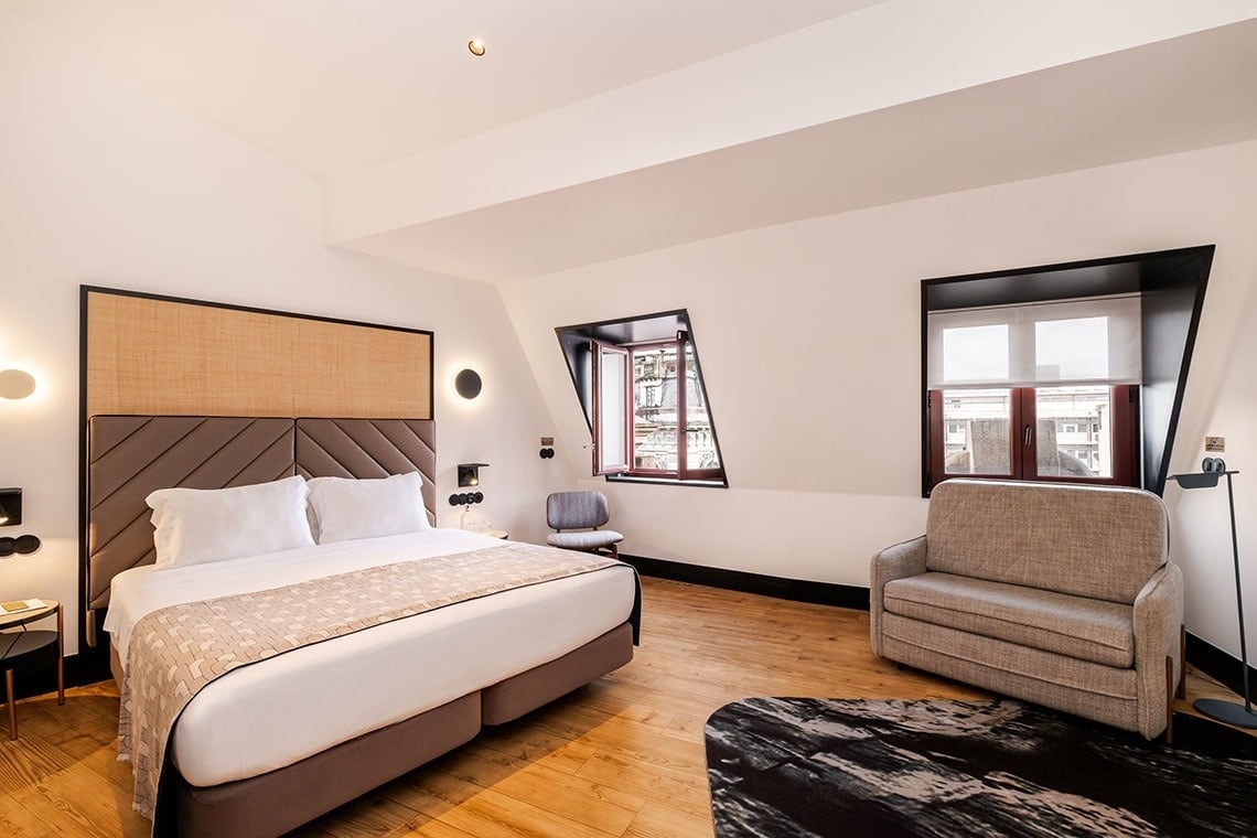 Um quarto contemporâneo em tons neutros, com cama grande, sofá, piso de madeira e duas janelas embutidas com molduras escuras.