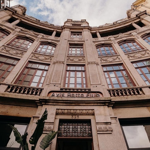 A low-angle shot captures the ornate, curved facade of a multi-story stone building with numerous windows, balconies, and decorative carvings, all under a cloudy sky.