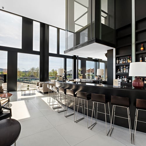 Um bar e lounge de hotel moderno e luminoso, com grandes janelas, um balcão preto elegante, assentos e um elevador de vidro no centro.