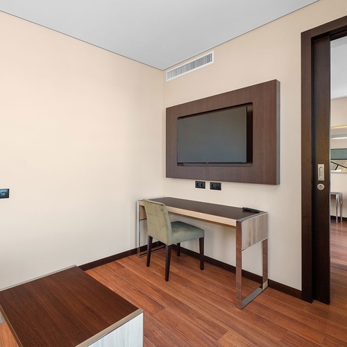 Um quarto de hotel moderno com paredes claras e piso de madeira, apresentando uma secretária, uma cadeira e uma televisão montada, com uma porta aberta revelando uma poltrona noutra divisão.