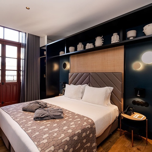 A modern and stylish hotel bedroom features a large bed with a patterned throw, dark blue walls, a built-in shelving unit, and natural light streaming through wooden French doors.