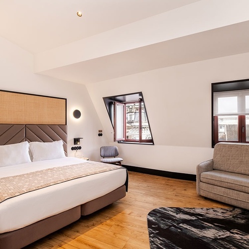 A brightly lit, modern hotel room features a large bed with a textured headboard, two dormer windows, a light gray sofa, and warm wooden floors.