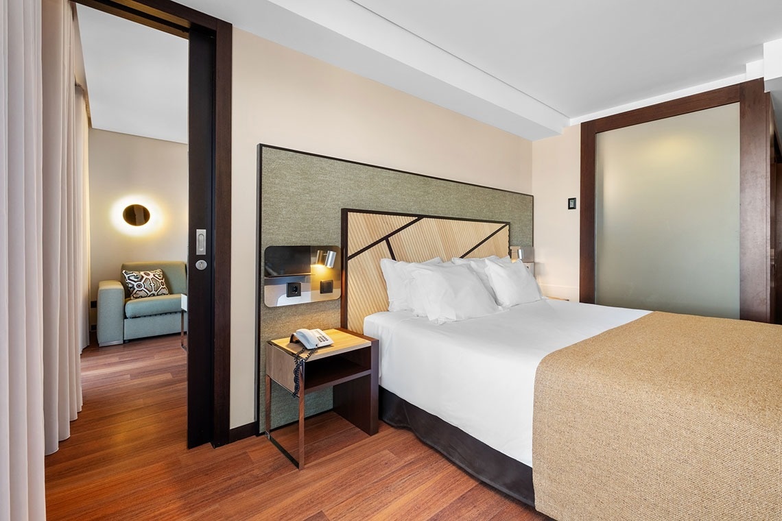 Um quarto de hotel moderno e bem iluminado apresenta uma cama arrumada, mesa de cabeceira e acesso a uma sala de estar adjacente.