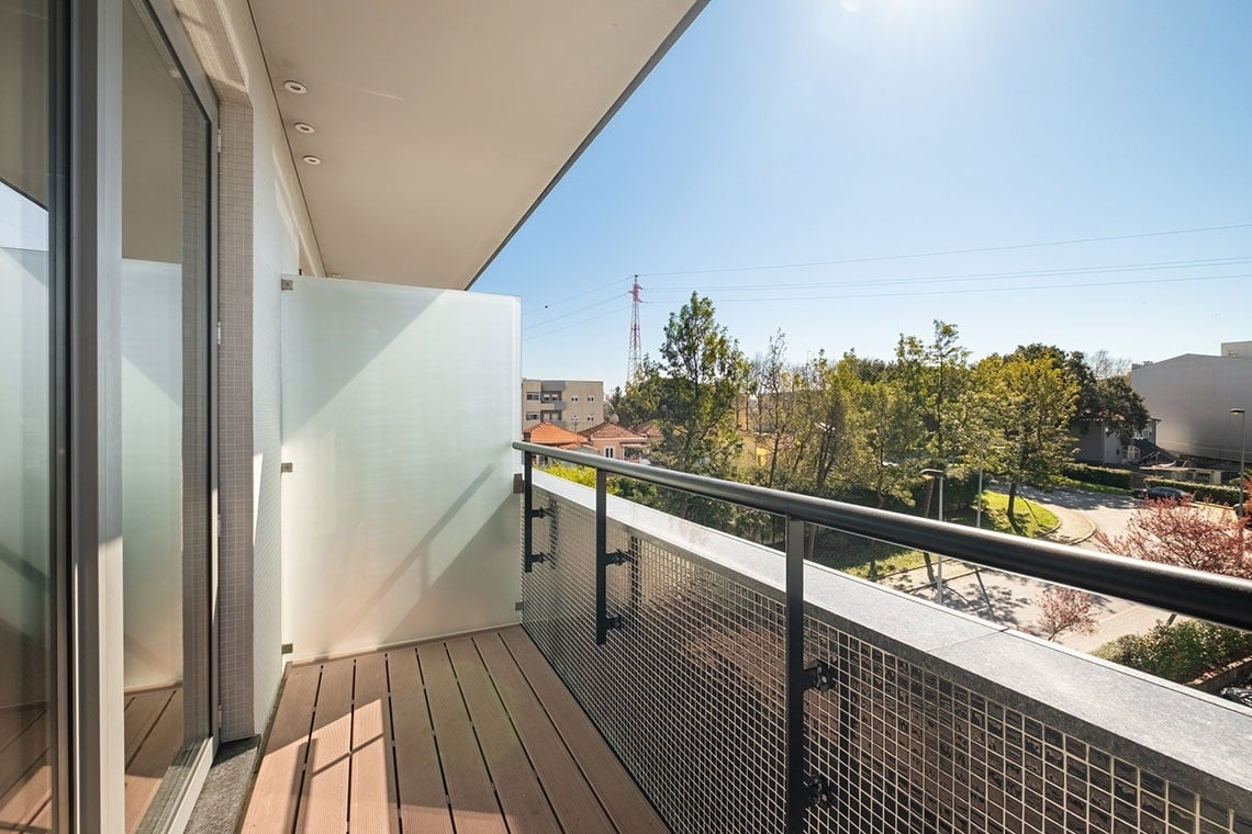 Moderna varanda com piso de madeira, painel de vidro fosco e grade metálica, oferecendo vista para uma paisagem urbana e arborizada sob um céu azul e ensolarado.