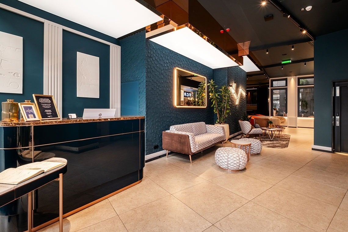 Um lobby de hotel moderno com balcão de recepção escuro e brilhante, paredes azul-petróleo texturizadas e uma área de estar aconchegante.